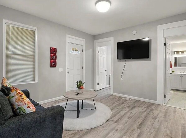 A modern living room with a grey couch, a round coffee table, a flat-screen TV mounted on the wall, and a wooden floor.