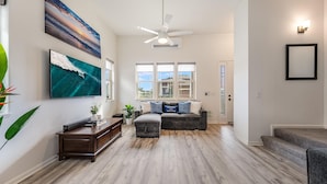 A spacious living room with hardwood flooring, a ceiling fan, a large flat-screen TV, a sectional sofa with decorative pillows, and a staircase leading to another level.