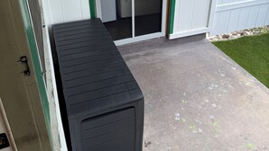 Un petit abri de jardin avec un banc, un lave-linge et une porte donnant sur une zone herbeuse.