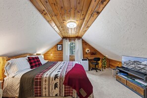A cozy attic bedroom with a wooden ceiling, a bed with patterned bedding, a desk with a computer, and a TV on the right. - Centennial Chalet , Tranquil cabin 20 min from Yellowstone (Island Park)