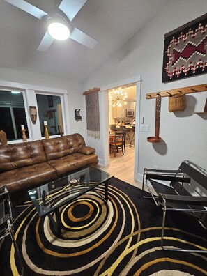 A living room with a brown leather sofa, a glass coffee table, a black and yellow patterned rug, and a modern metal chair. - Modern Tiny House w/ Heated Pool in Kitty Hawk (Kitty Hawk)