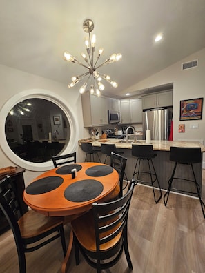 A modern kitchen with a central island, stainless steel appliances, and a dining area with a round table and black chairs. - Modern Tiny House w/ Heated Pool in Kitty Hawk (Kitty Hawk)