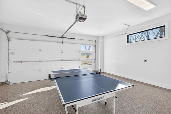 A ping pong table in a room with a garage door, a projector, and a window with a view of trees.