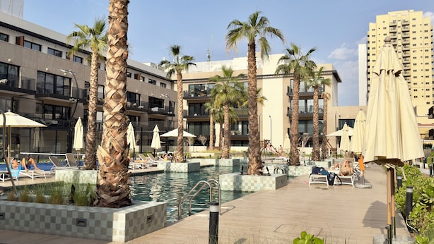 A pool area with lounge chairs and umbrellas, surrounded by palm trees and modern buildings. - Modern Studio in Oakley Square JVC with Pool, Gym & Cinema (Dubai)