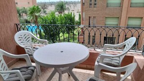 A balcony with a round table and four white chairs, a metal railing, and a view of a swimming pool and greenery. - Apartment Gueliz - Near ONCF station - Marrakech (Marrakech)