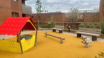 Een speeltuin met een gele speelconstructie, bankjes en een zandbak.