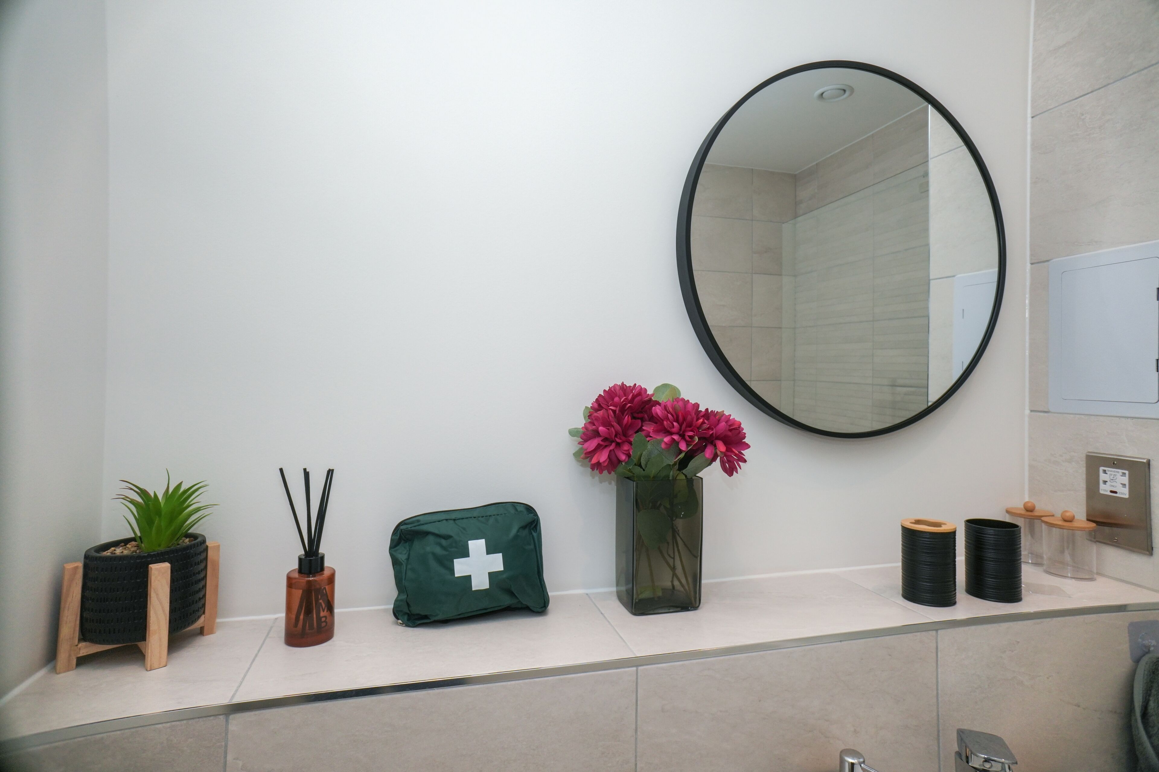 Une salle de bain avec un miroir, une plante, un sac vert et un vase rempli de fleurs.