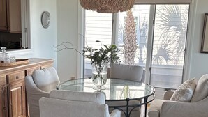 Ein modernes Wohnzimmer mit einem runden Glastisch, zwei weißen Sesseln, einer gewebten Pendelleuchte und einem großen Fenster mit Blick auf Palmen.