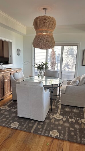 Een moderne woonkamer met een ronde glazen tafel, twee witte fauteuils, een geweven hanglamp en een groot raam met uitzicht op palmbomen.