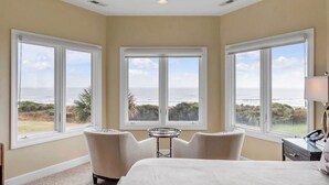 Ein Hotelzimmer mit einem großen Bett, zwei Sesseln, einem kleinen Tisch und drei großen Fenstern mit Blick aufs Meer.