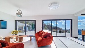 Un vaste espace de vie comprenant un canapé rouge, une table basse en bois et un fauteuil jaune. De grandes portes-fenêtres coulissantes donnent accès à une terrasse extérieure offrant une vue sur le paysage.