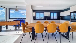Un espace repas moderne avec une table en bois et des chaises jaunes, un baby-foot et une vue sur le ciel grâce à de grandes fenêtres.