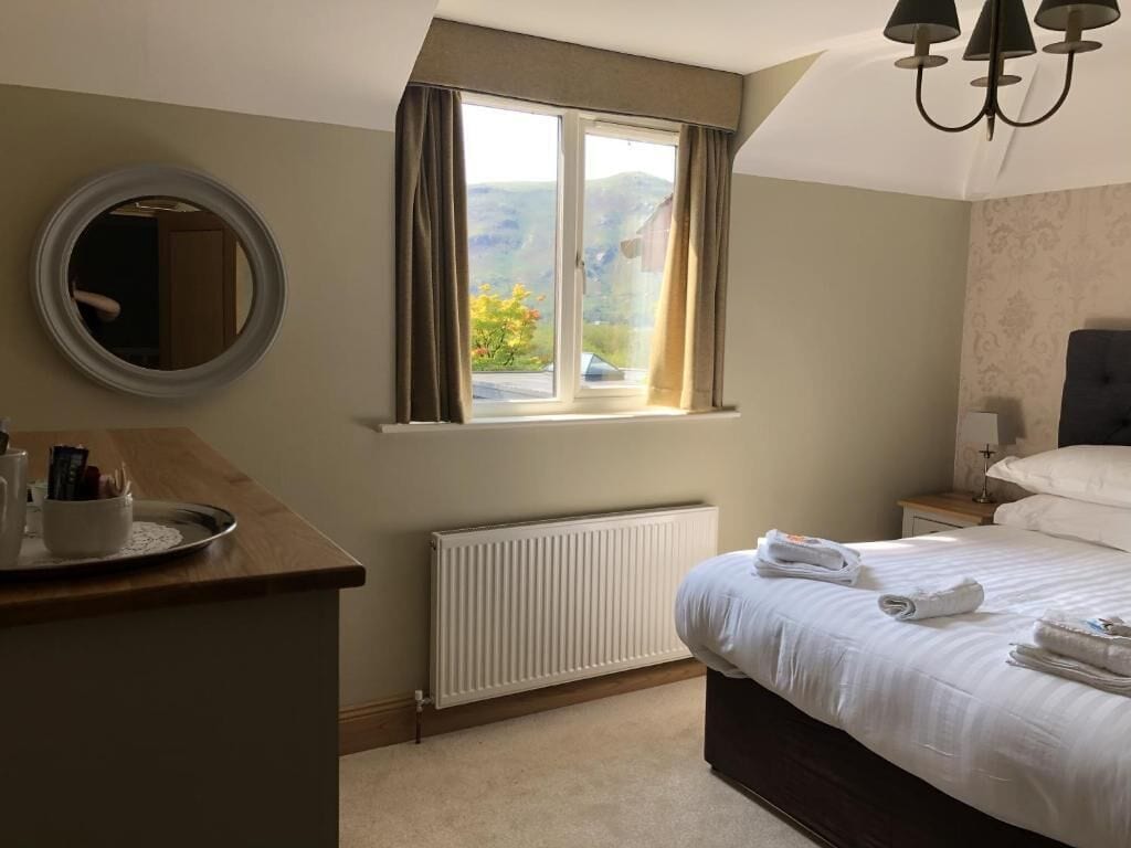 A bedroom with a bed, a bedside table, a mirror, a window with curtains, a radiator, and a chandelier.