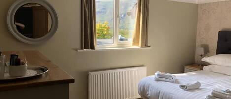 A bedroom with a bed, a bedside table, a mirror, a window with curtains, a radiator, and a chandelier.