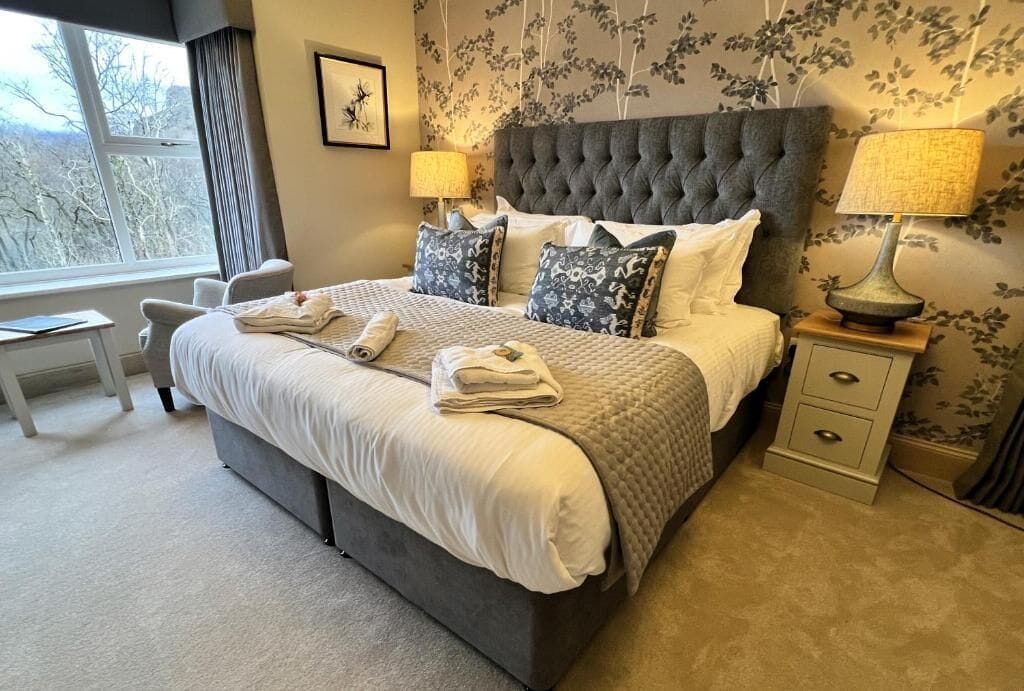 Une chambre à coucher avec un grand lit, deux tables de chevet avec des lampes, une chaise, une petite table et une fenêtre donnant sur un beau paysage.