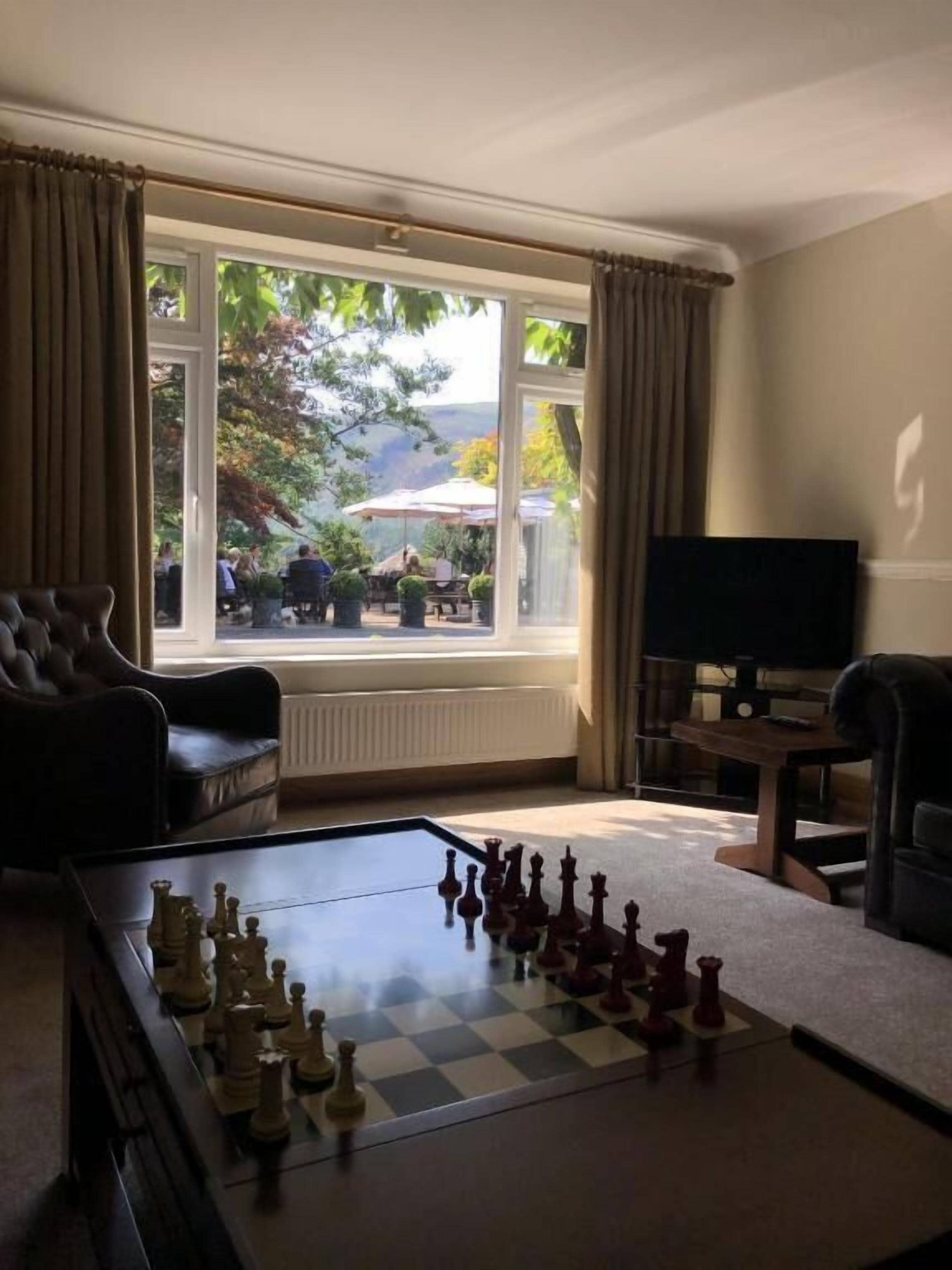 A living room with a chessboard, a sofa, a television, and a window with a view of outdoor seating.