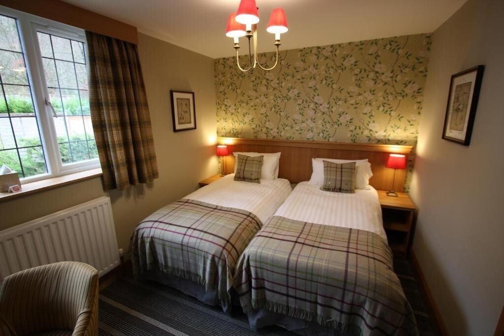 A double room with two beds, a window with curtains, a radiator, a chair, and a wall with floral wallpaper.