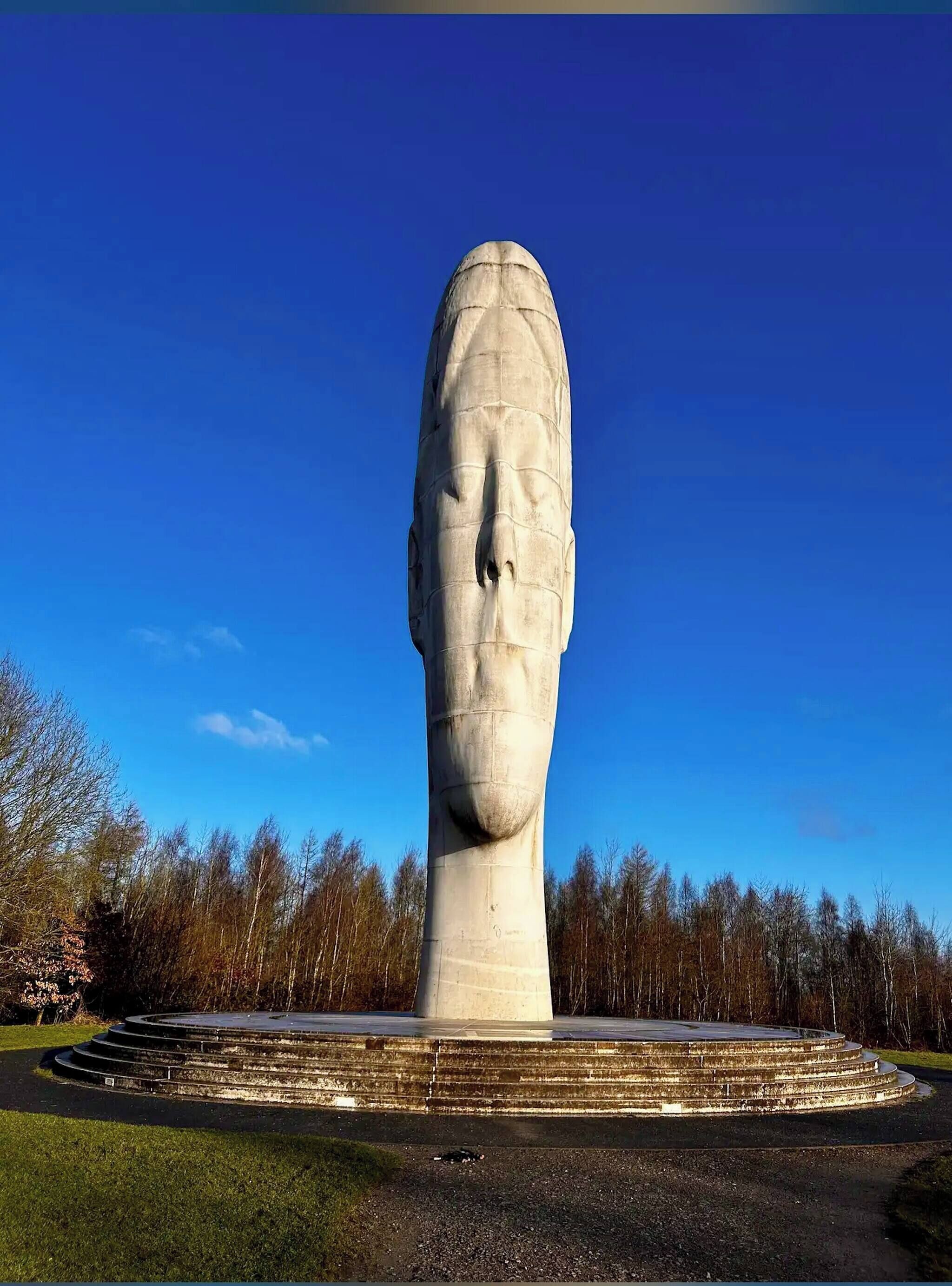 En stor stenskulptur som föreställer ett mänskligt huvud, med en cirkelformad bas och trappsteg som leder upp till den. Skulpturen är placerad i en park med träd i bakgrunden.