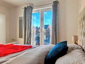 A bedroom with a bed, a window with curtains, a view of buildings, and a lamp on a bedside table. - Contractor-friendly 2BR Gem Wi-fi & Free Parking (Warrington)