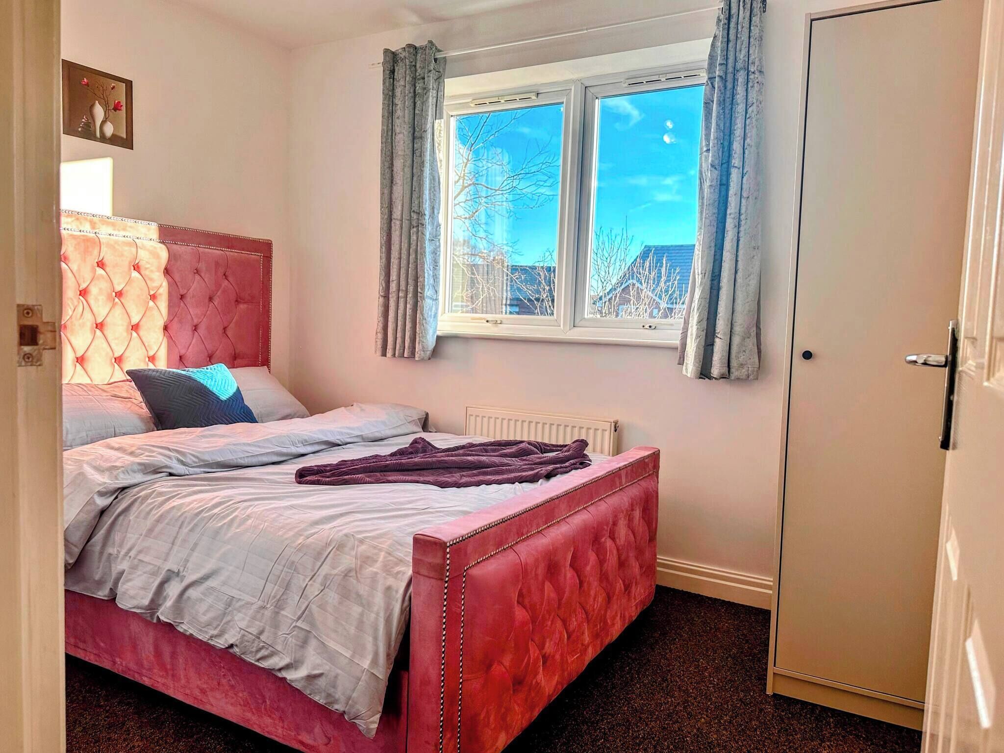 Une chambre avec une tête de lit rose à motifs capitonnés, un lit recouvert d’une couette blanche et grise, un plaid violet et une fenêtre avec des rideaux.