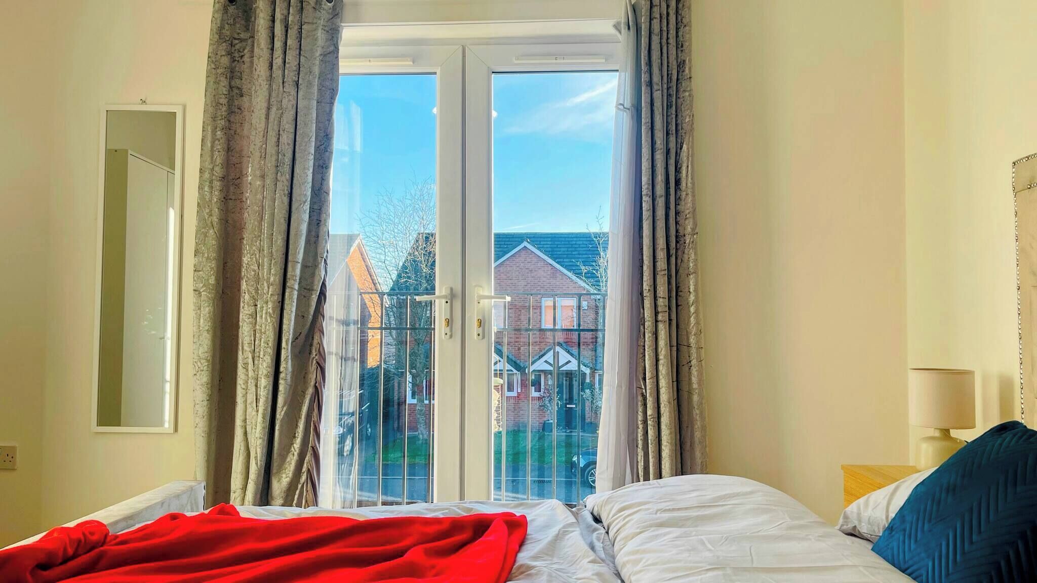 A bedroom with a bed, a window with curtains, a view of houses, and a lamp on the nightstand.