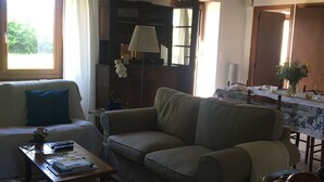 A living room with a sofa, a coffee table, and a dining table with chairs. There is a window with curtains, a lamp, and a vase with flowers. - Delightful 2-bedroom cottage perfect for a revitalizing retreat in Brittany (Ruffiac)