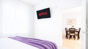Quarto de hotel com cama, TV fixada na parede exibindo 'NETFLIX', área de refeições com mesa e cadeiras, e porta que dá acesso a outro cômodo.