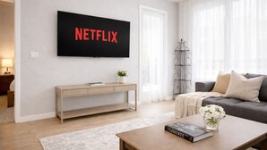 Uma sala de estar moderna com uma televisão de tela plana exibindo 'NETFLIX', um sofá, uma mesa de centro com um livro e um aparador com uma planta.