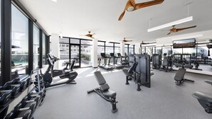Uma academia moderna com ventiladores de teto, diversos equipamentos de exercício e amplas janelas com vista para a cidade.