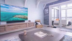 Ett modernt vardagsrum med en stor platt-TV, en korg av rotting, ett soffbord och en skjutdörr i glas som leder ut till en utomhusplats med sittmöbler.