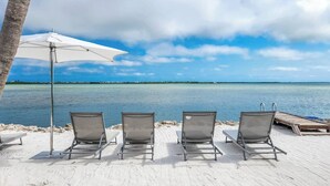 En avslappnad lounge vid stranden med tre solstolar, ett vitt parasoll och en träbrygga som sträcker sig ut i vattnet.