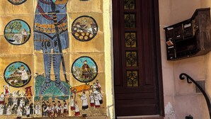 Eine Gebäudefassade mit einem großflächigen Mosaikbild, das historische Szenen und Figuren darstellt, einer dunklen Holztür und einem kleinen, schwarzen Briefkasten.