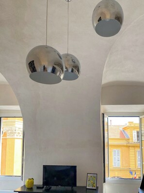 A room with a modern chandelier, a flat-screen TV, and a window with a view of yellow buildings. - Chic Ferrié Riviera apartment (Menton)