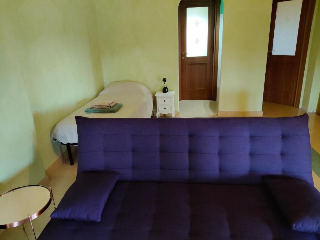 A room with a dark blue sofa, a small round table, a bed with a white headboard, a nightstand with a lamp, and a door with a frosted glass panel.