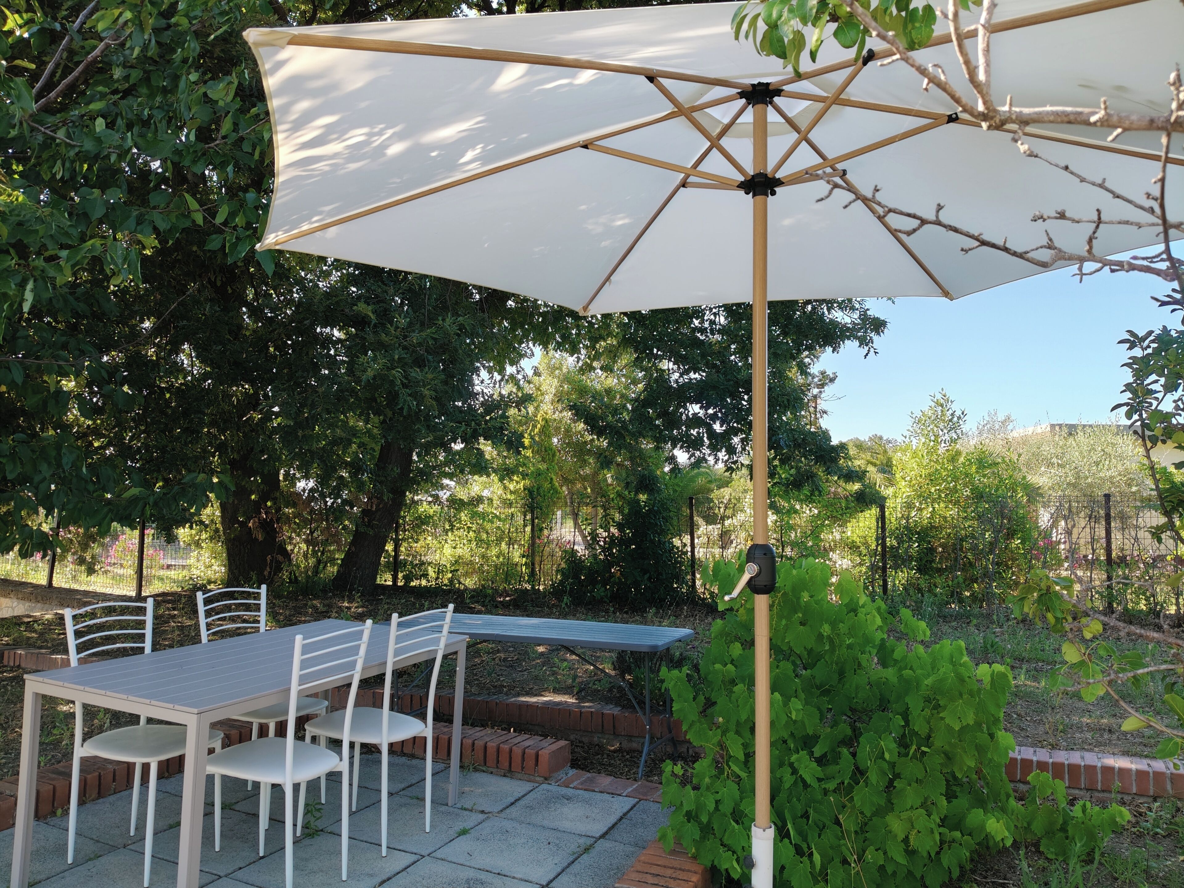 An outdoor patio with a table and chairs, a white umbrella, and a garden with trees and plants.