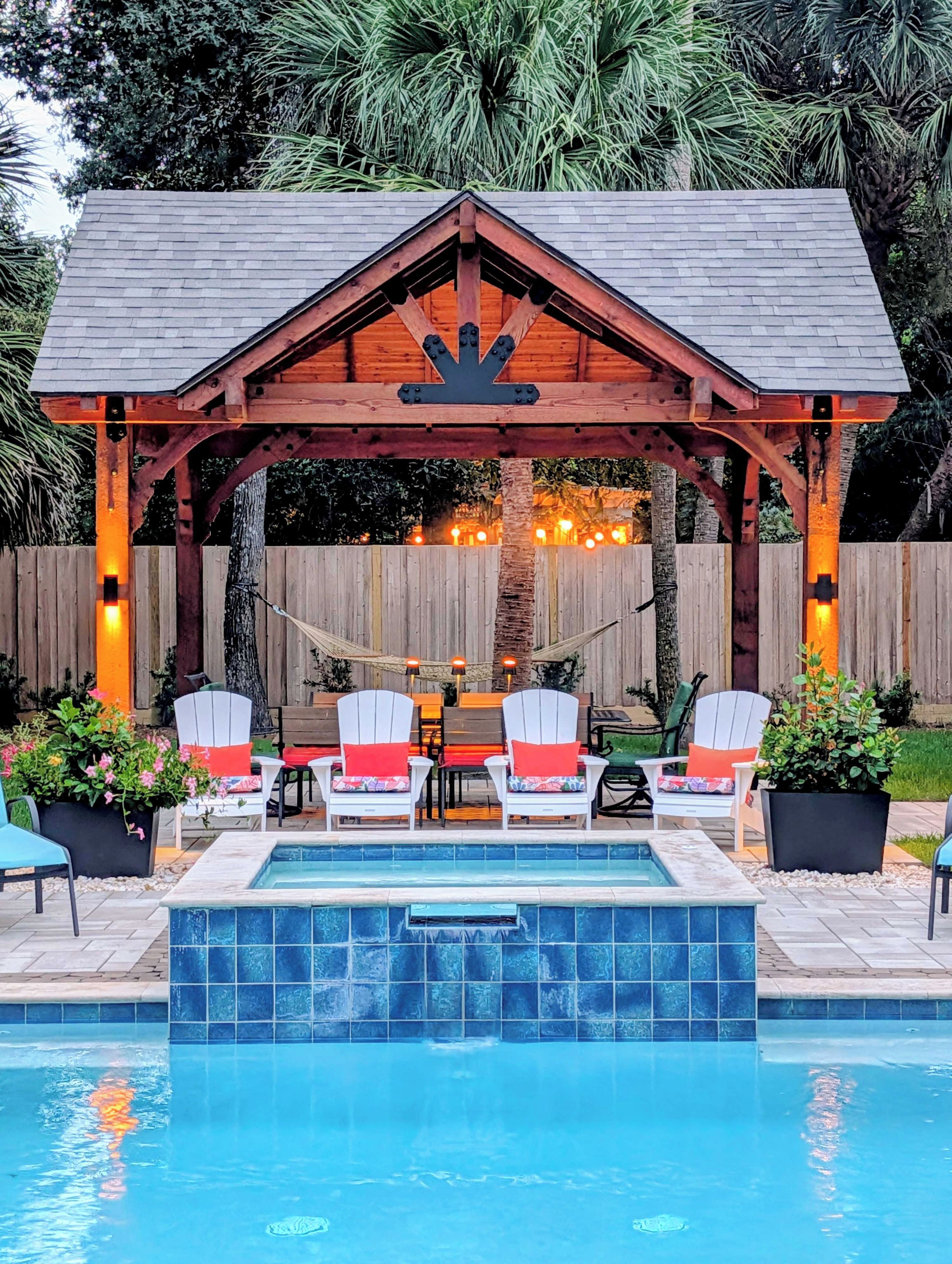 A wooden gazebo with a pool table and chairs, surrounded by a wooden fence and potted plants.