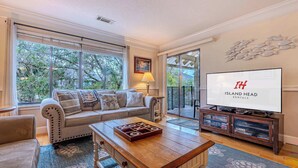 A living room with a sofa, coffee table, and TV. There is a large window with blinds, a door leading to a balcony, and a wall-mounted TV displaying "Island Head Rentals". - 2BR Lagoon View Villa - Walk to Coligny (Hilton Head Island)