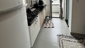 Et lille køkken med et køleskab, komfur og mikroovn. Der er en lille måtte på gulvet, og et vindue med gardiner er synligt.
