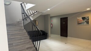 Una escalera con barandilla negra, un extintor y una puerta marcada con el número 108 en un pasillo con suelo de baldosas.
