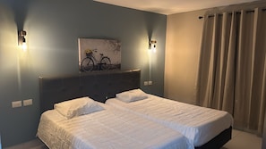 Un lit double avec du linge de lit blanc, une tête de lit, deux appliques murales et un tableau représentant un vélo.