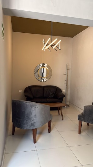 Un salon moderne avec un canapé en cuir, deux fauteuils gris, un miroir rond et un luminaire au plafond au design original.