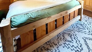 Ein Holzbett in einer Hütte mit schräg laufender Decke und einer Tür im Hintergrund.