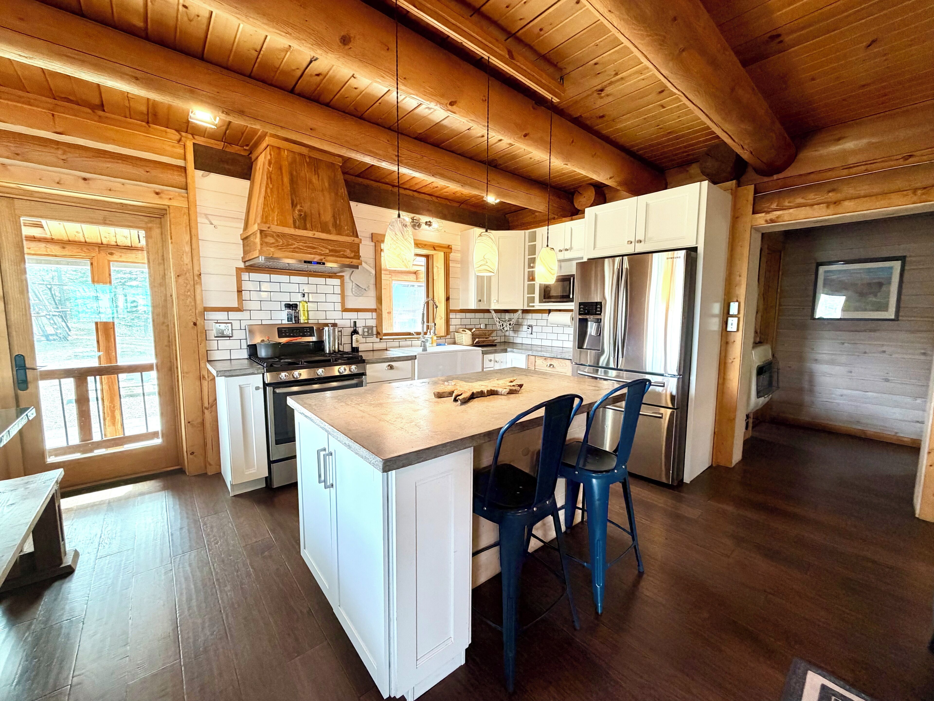 A modern kitchen with wooden beams, white cabinetry, stainless steel appliances, and a central island with two blue chairs.