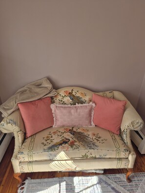 En sofa i vintage-stil med blomstret betræk og dekorative puder, placeret på et trægulv med et mønstret tæppe.