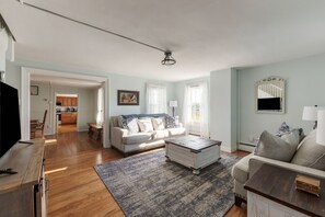 A living room with a sofa, coffee table, and a TV. There is a kitchen in the background.