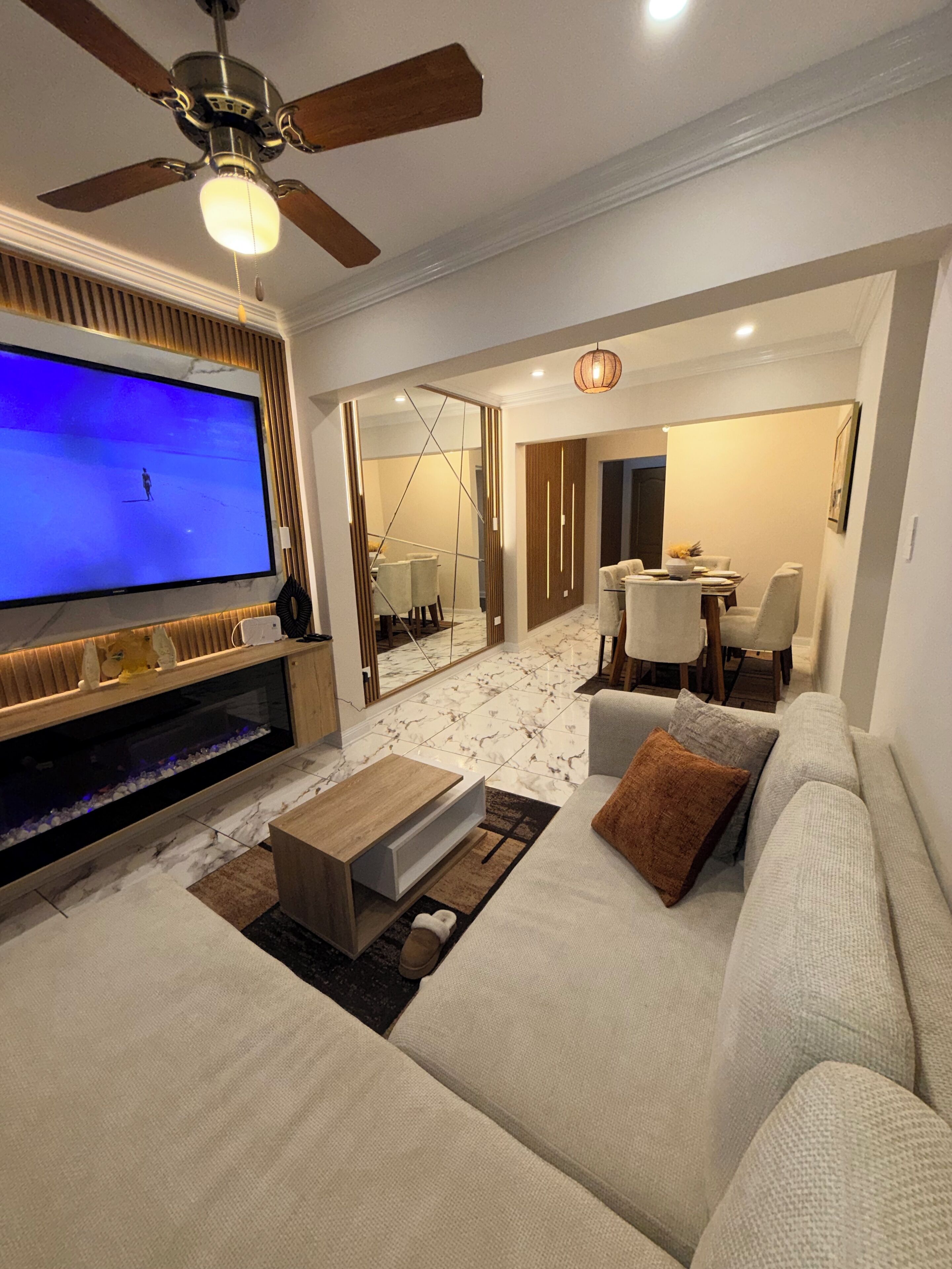 A modern living room with a flat-screen TV, a ceiling fan, a sofa, a coffee table, and a dining area with chairs and a table.