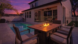 Un área de piscina moderna al aire libre con una mesa y sillas de madera, un hogar para fuego y una zona de estar cubierta.