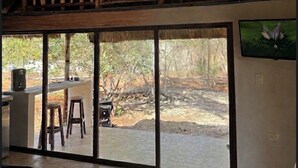 Een kamer met rieten dak, een balkon met houten leuningen, een eethoek met krukken, en grote schuifglazen deuren die leiden naar een buitenruimte.