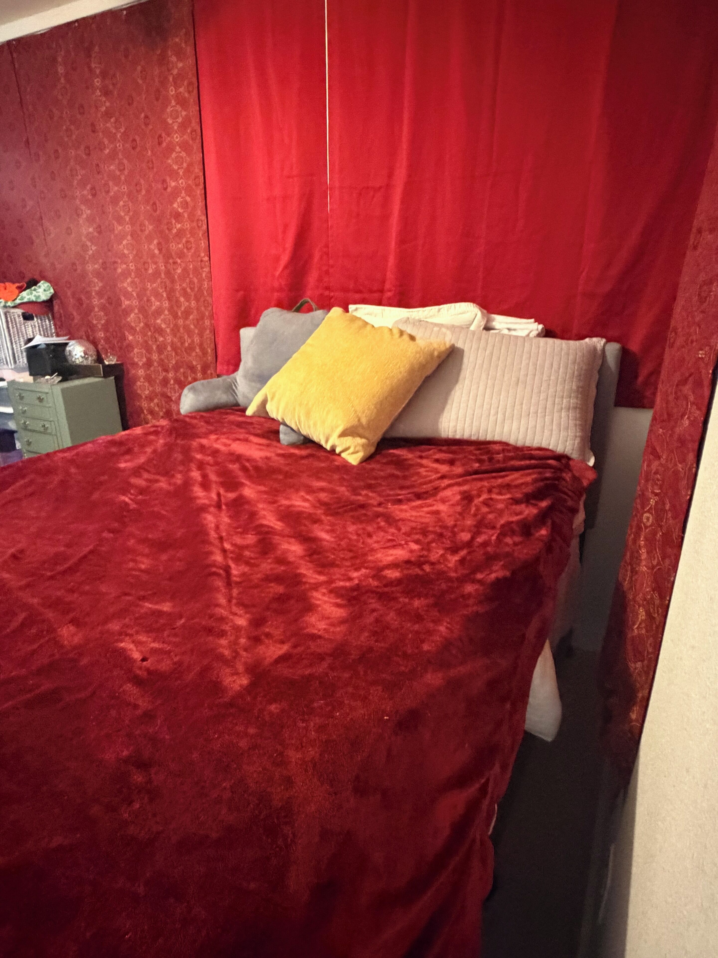 A bed with a red velvet bedspread, a yellow pillow, and a gray pillow. There is a green dresser with a mirror and a small table with a lamp.