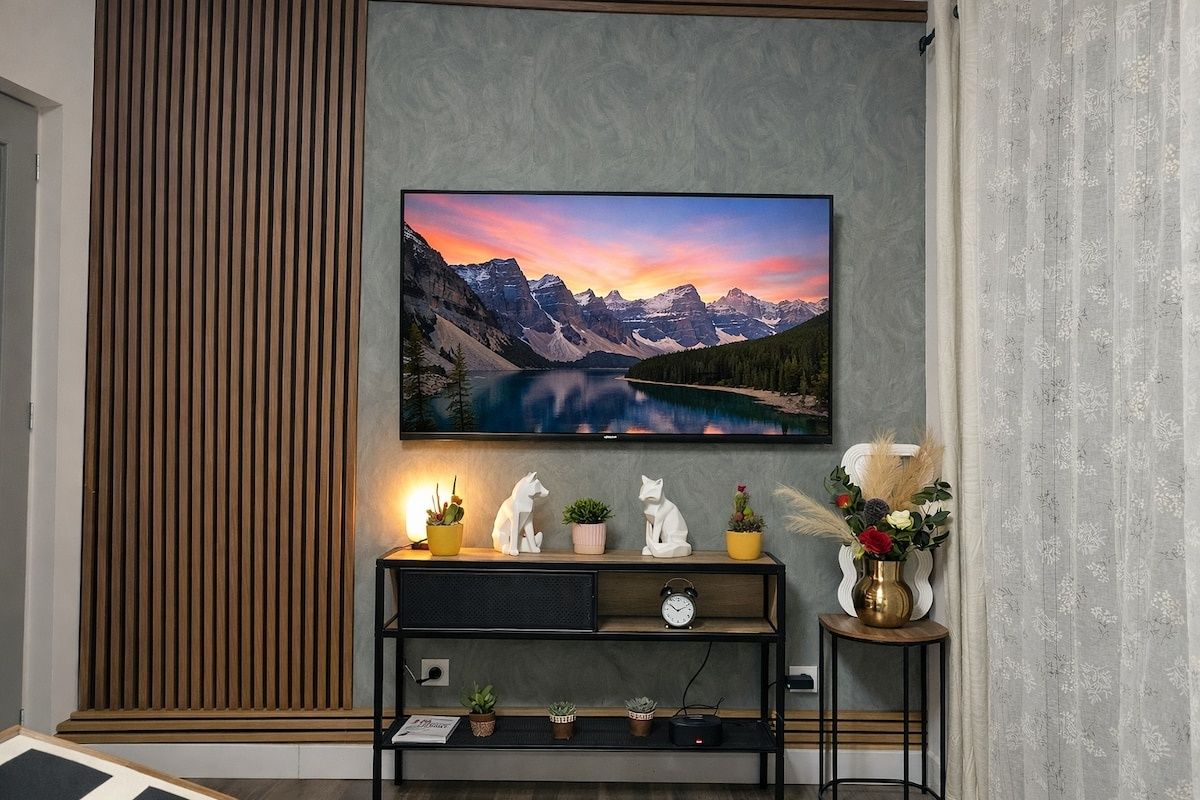 A television mounted on a wall with a scenic landscape image, a wooden panel, a black entertainment unit with decorative items, and a curtain.